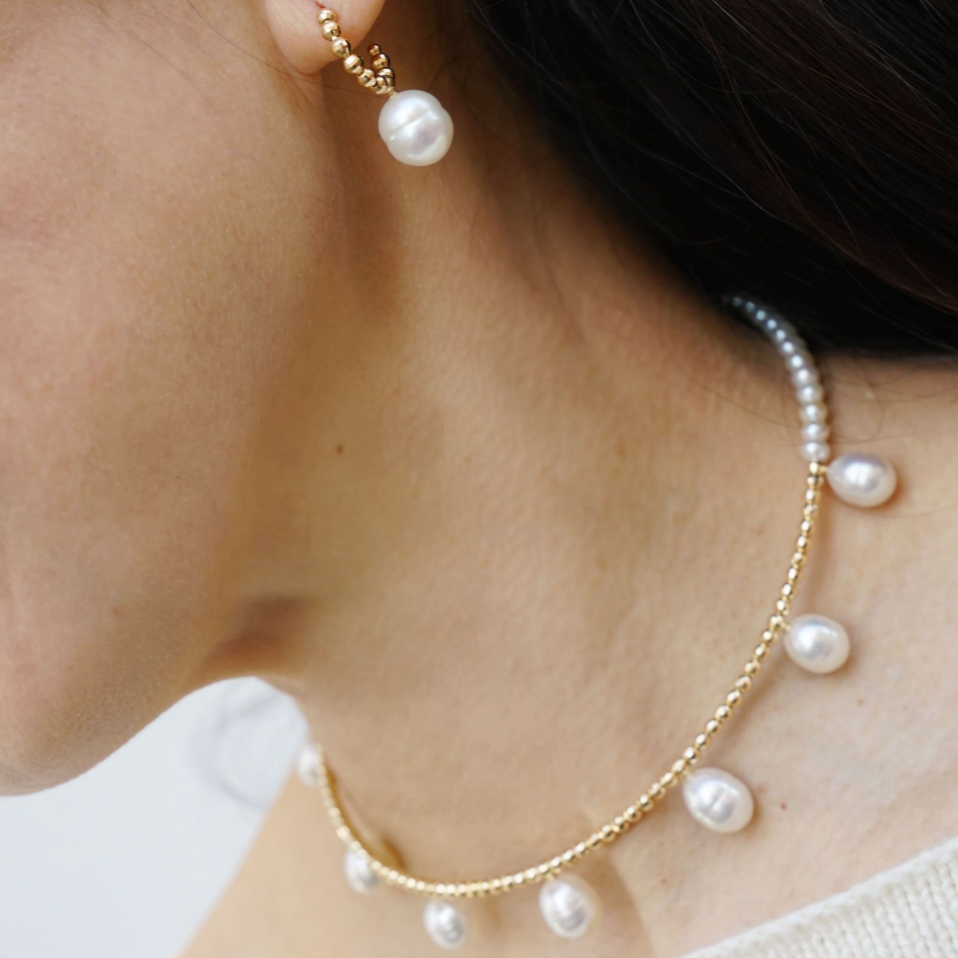 Elevate your style with a woman exuding grace, wearing a pearl necklace and earrings. Experience the allure of Pearl Dot x Gold Piercing.