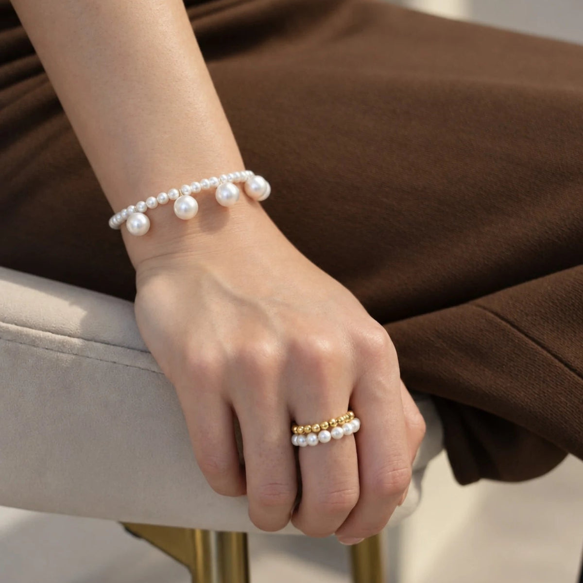 Blair Bracelet on a wrist resting on a chair arm, larger freshwater pearls hanging from a row of small pearls, worn with a gold bead and pearl ring