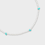 Close-up of white pearl necklace featuring turquoise enamel heart charms with silver-tone edges on a soft gray background