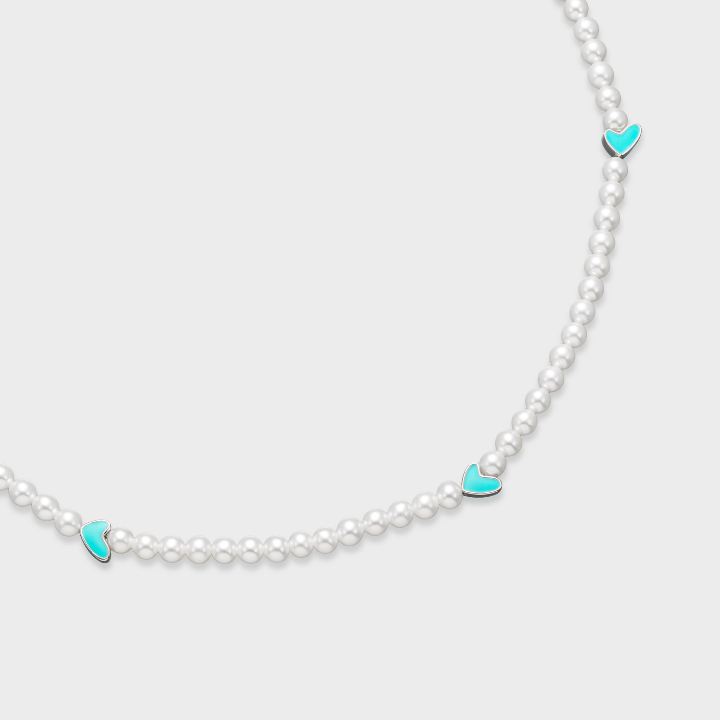 Close-up of white pearl necklace featuring turquoise enamel heart charms with silver-tone edges on a soft gray background
