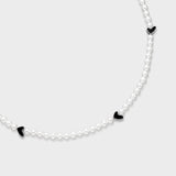 Close-up of white pearl necklace featuring black enamel heart charms with silver-tone edges on a soft gray background