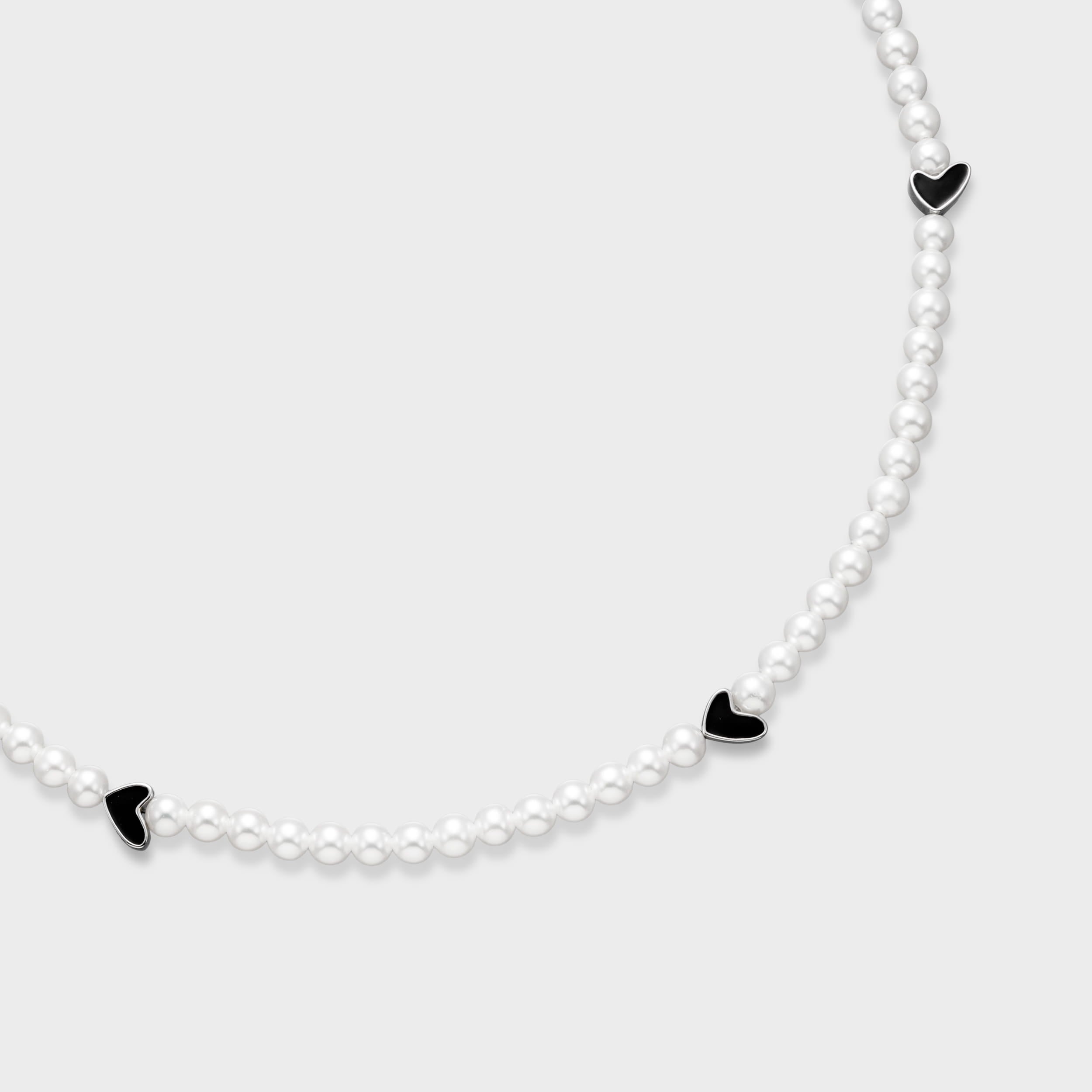 Close-up of white pearl necklace featuring black enamel heart charms with silver-tone edges on a soft gray background