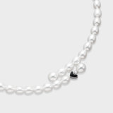 Close up of white oval freshwater pearl necklace with small black enamel heart charm accent on a light gray background.